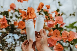 a person holding two tubes of lotion in front of orange flowers