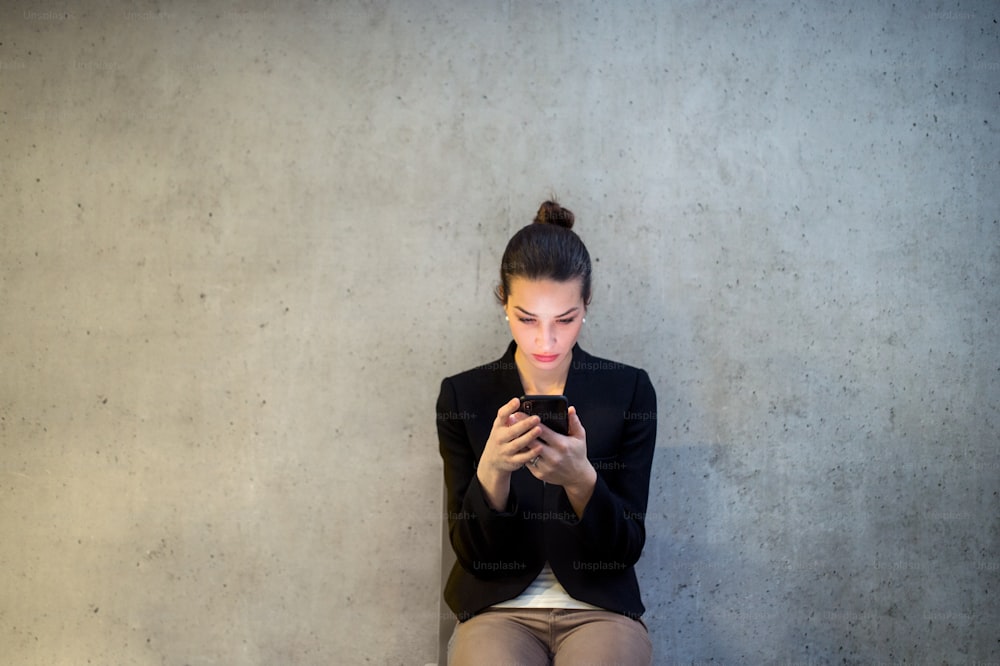 A front view of happy young business woman with smartphone sitting against concrete wall in office.
