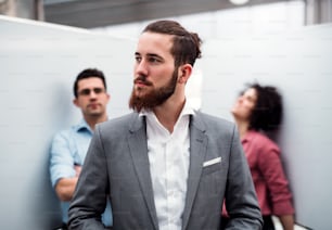 A portrait of young businessman in suit standing in office, colleagues in the background.