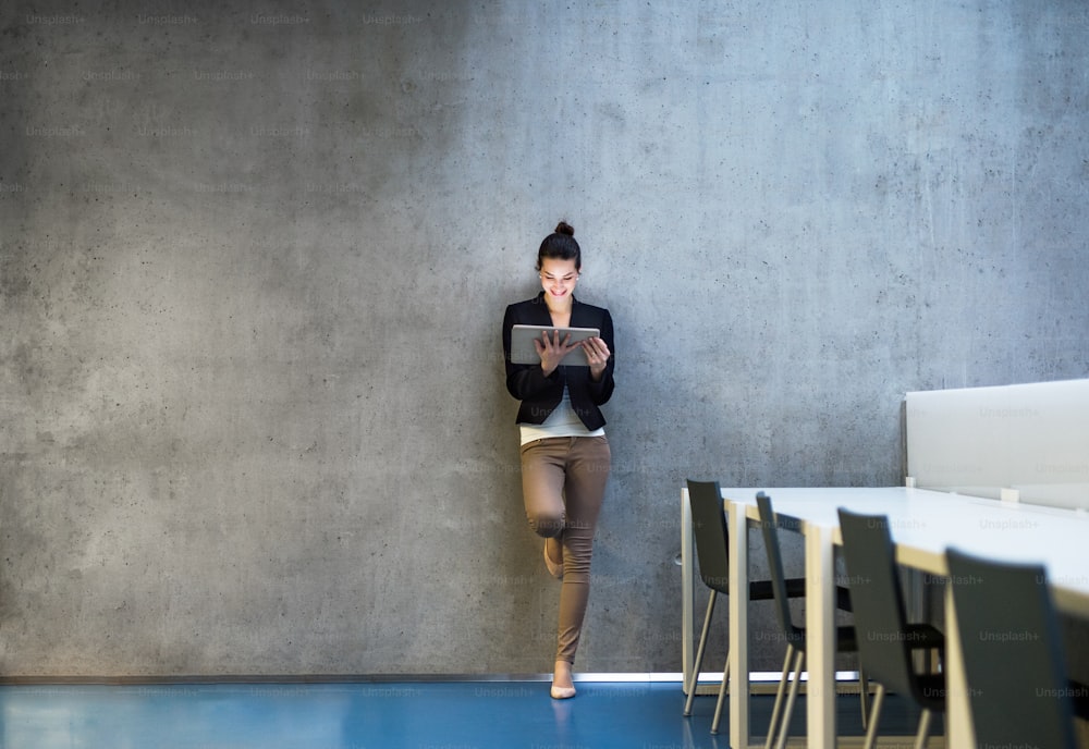 A portrait of happy young business woman with digital tablet standing against concrete wall in office.