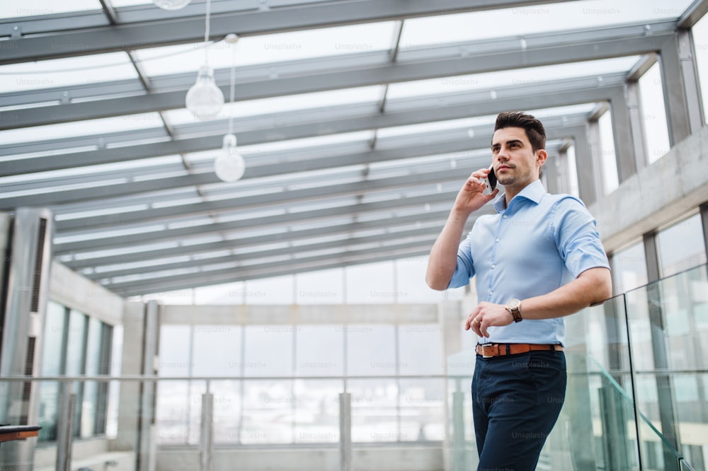 Ein Blick aus einem niedrigen Winkel auf einen jungen, ernsthaften Geschäftsmann mit Smartphone auf dem Flur vor dem Büro, der einen Anruf tätigt.
