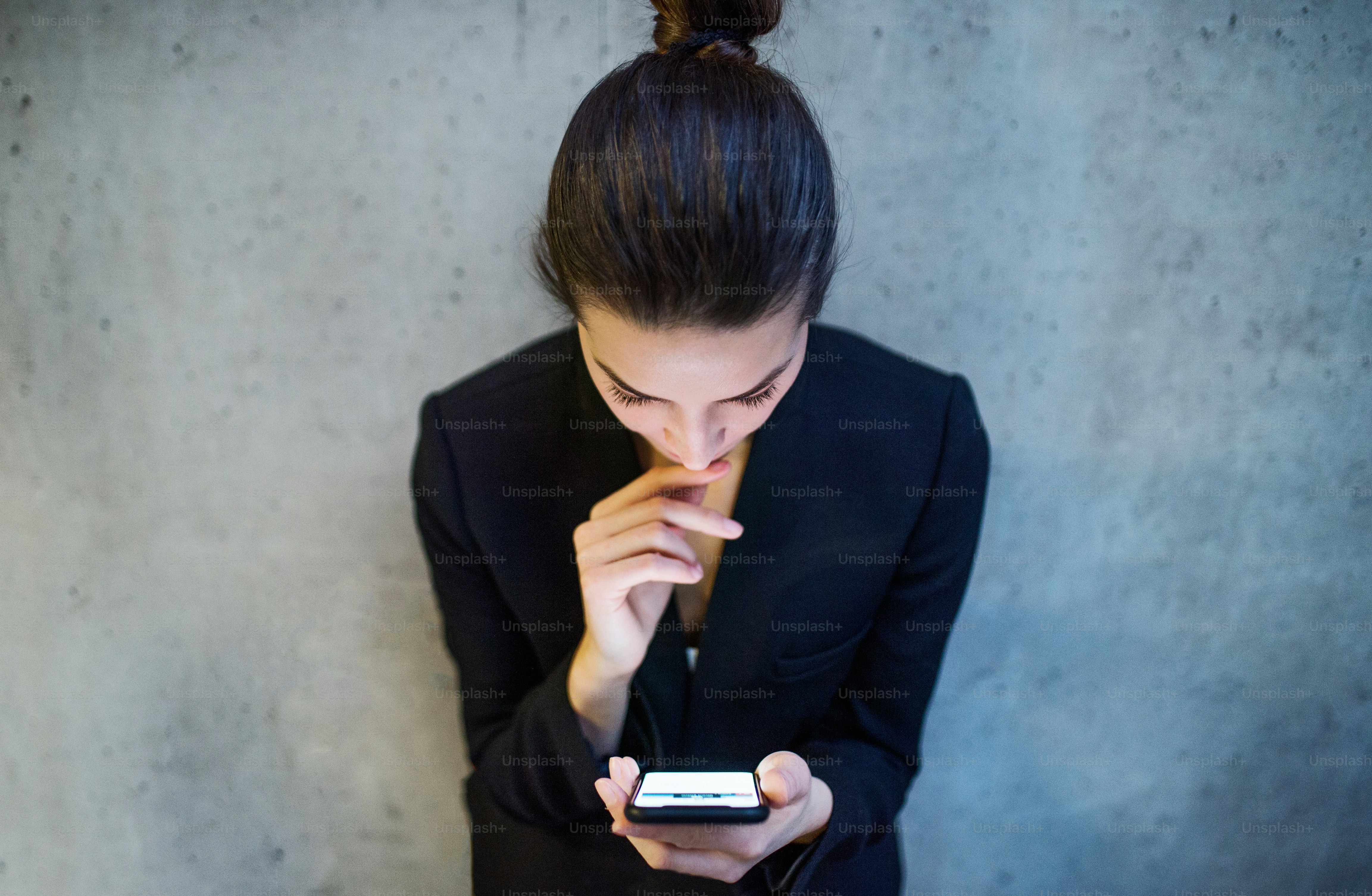 A top view of happy young business woman with smartphone standing against concrete wall in office.