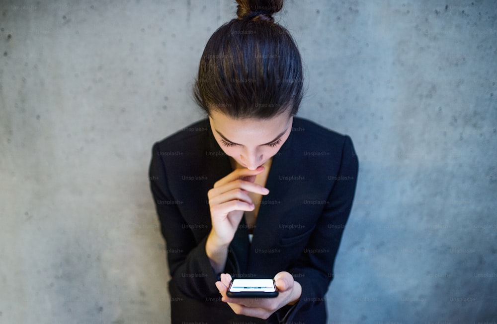 A top view of happy young business woman with smartphone standing against concrete wall in office.