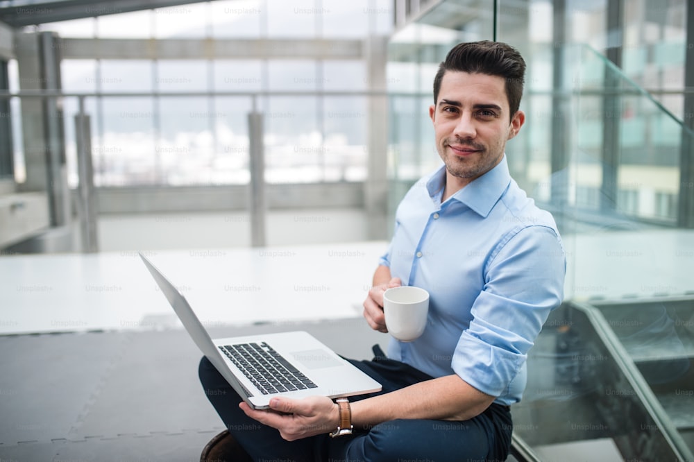 Um retrato de um jovem empresário com laptop e café sentado no chão no corredor do lado de fora do escritório.