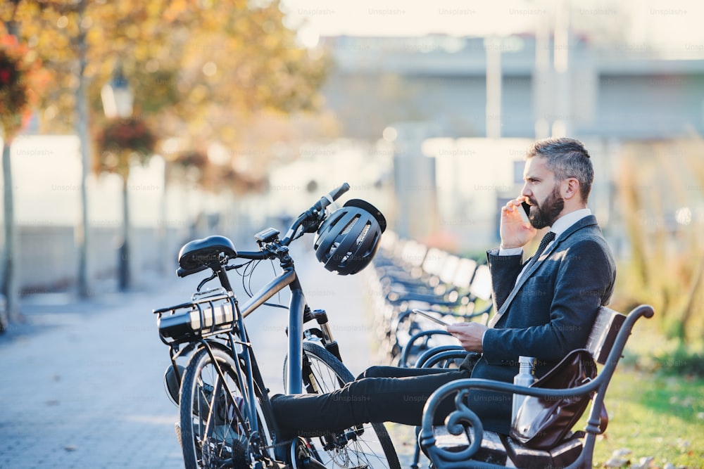Una vista lateral del viajero de negocios con teléfono inteligente y bicicleta sentado en el banco de la ciudad, haciendo una llamada telefónica.