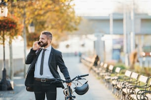A businessman commuter with bicycle walking home from work in city, using smartphone.
