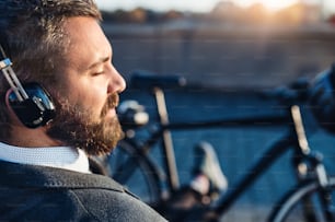 A close-up of businessman commuter with headphones and bicycle sitting on bench in city, listening to music. Copy space.