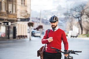 Pendolare uomo d'affari hipster con la bicicletta sulla strada per lavorare in città, in piedi e usando lo smartphone.