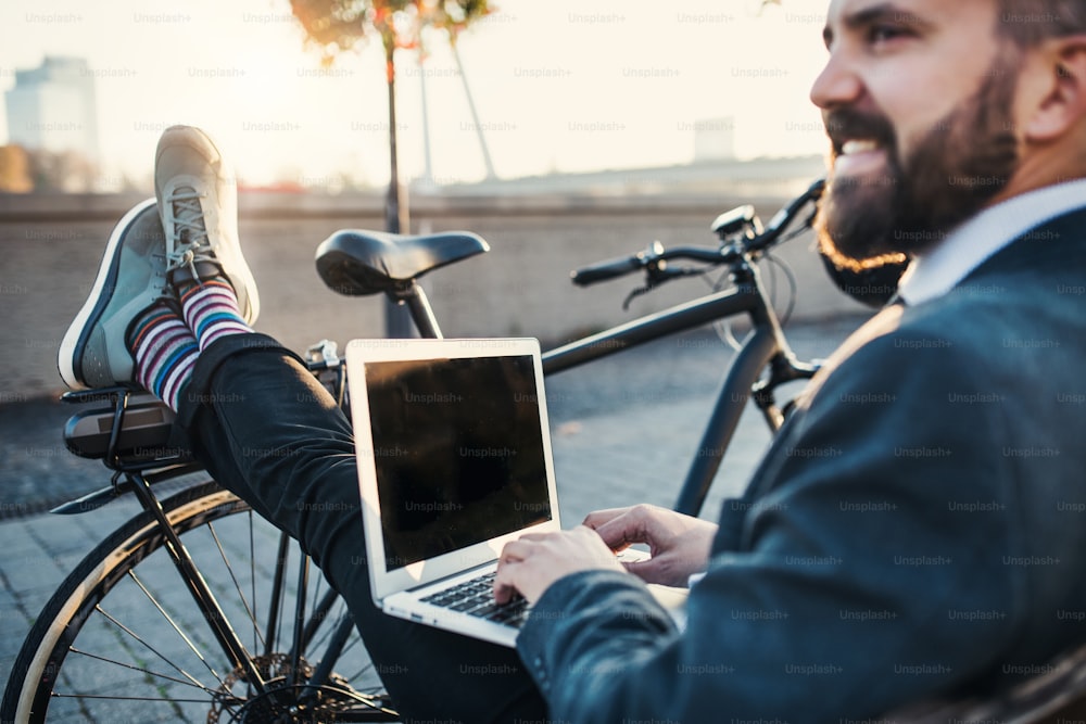 Ein Geschäftsmann, Pendler mit Fahrrad, sitzt auf einer Bank in der Stadt und benutzt einen Laptop, wenn er von der Arbeit nach Hause fährt. Speicherplatz kopieren.