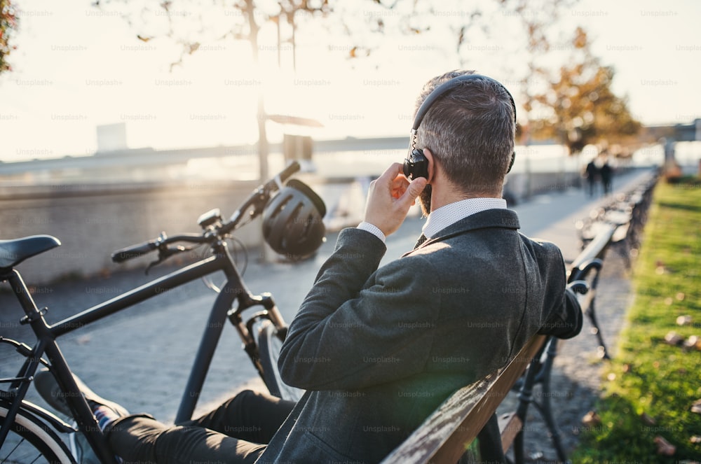 A rear view of businessman commuter with headphones and bicycle sitting on bench in city, listening to music.