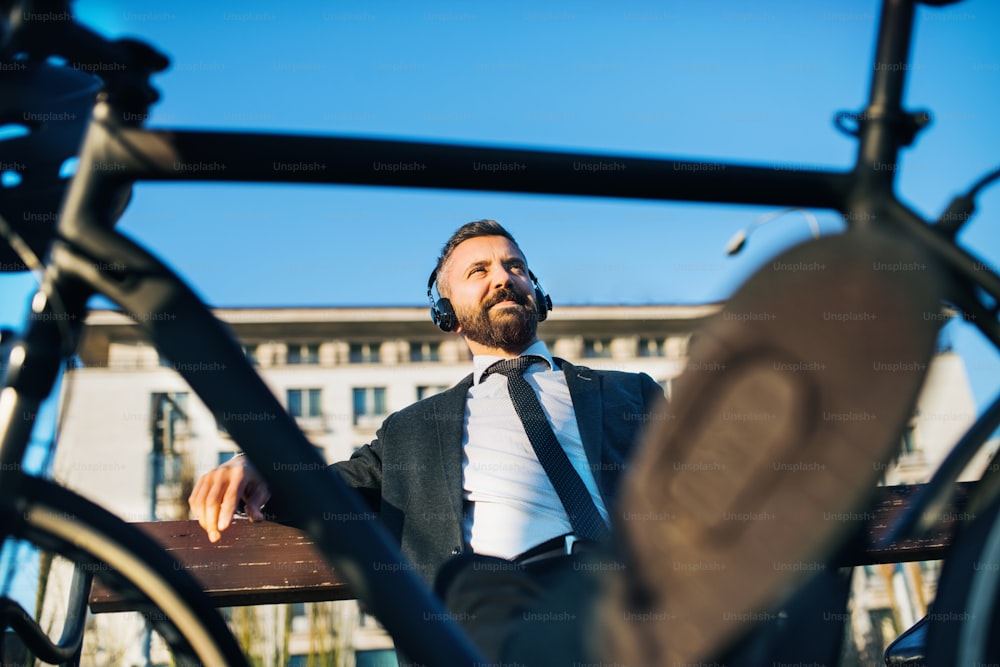 Businessman commuter with headphones and bicycle sitting on bench in city, listening to music.