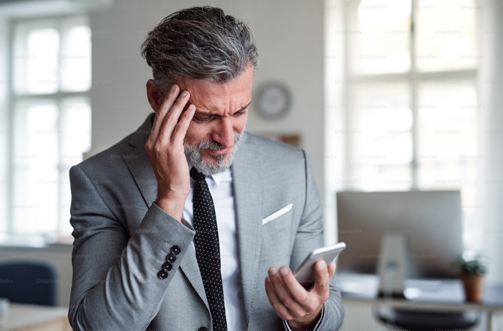 Un hombre de negocios triste y frustrado con un teléfono inteligente parado en una oficina, leyendo malas noticias.