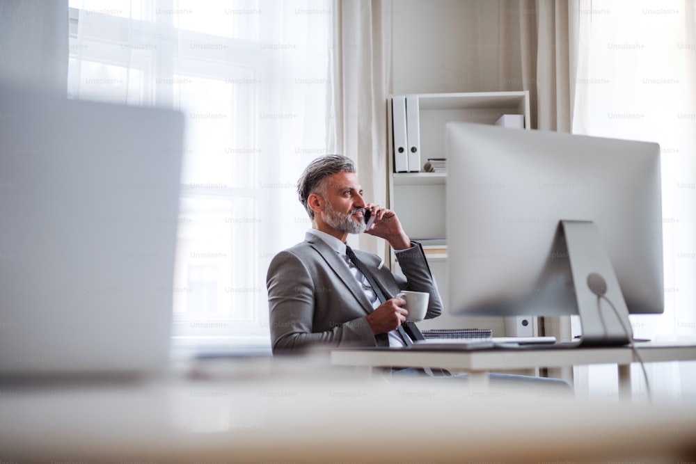 Ernsthafter reifer Geschäftsmann mit Smartphone, Kaffee und Desktop-Computer, der in einem Büro am Tisch sitzt und telefoniert.