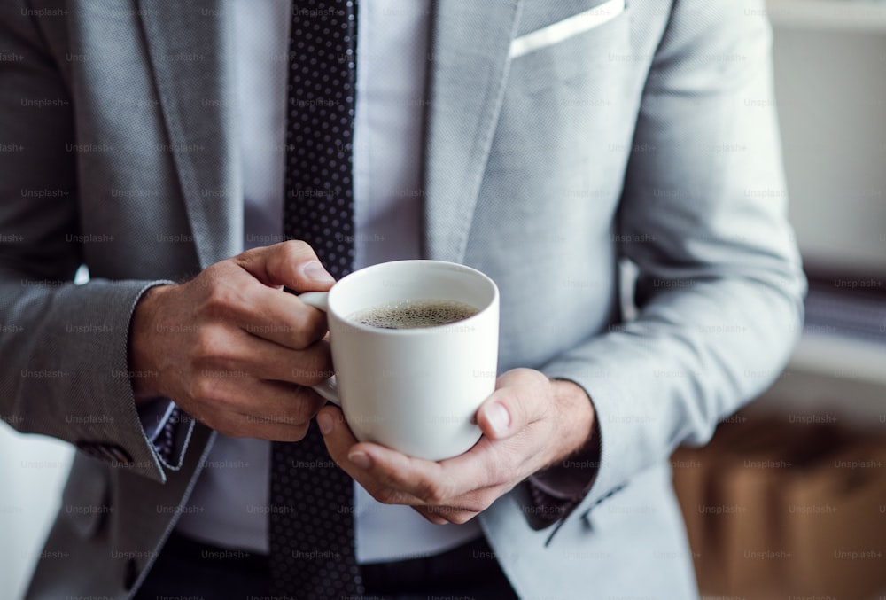 A midsection of businessman stabding in an office, holding a cup of coffee. A close-up.