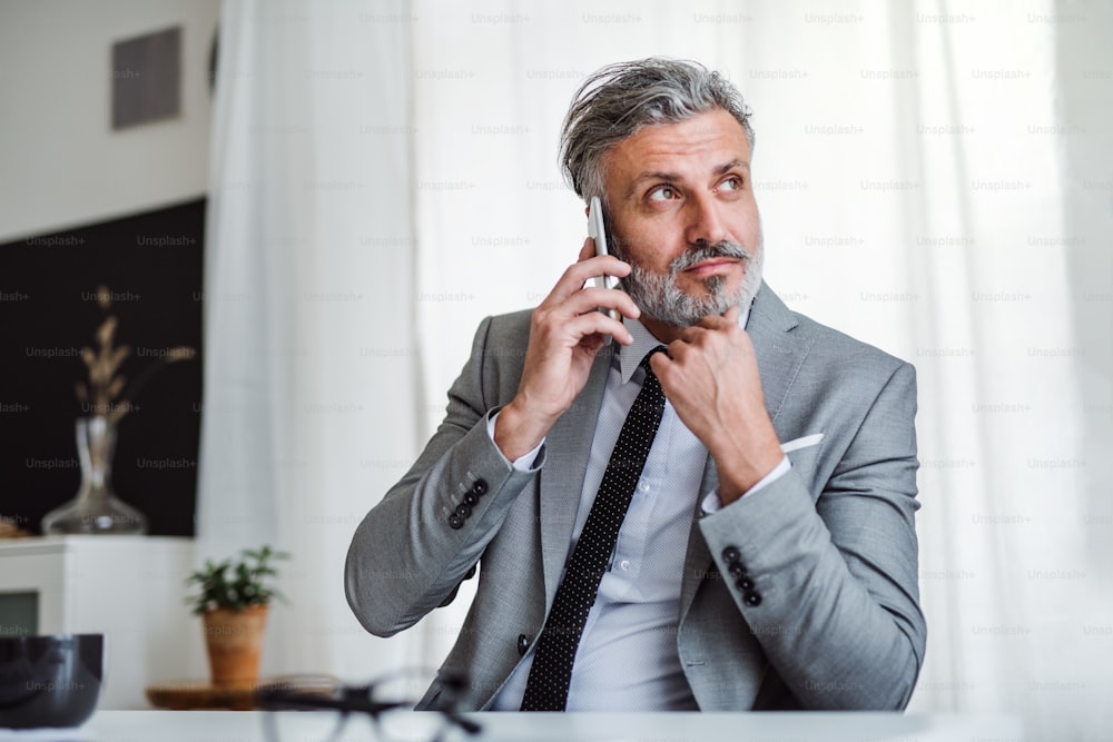 A serious mature businessman with smartphone sitting at the table in an office, making a phone call.