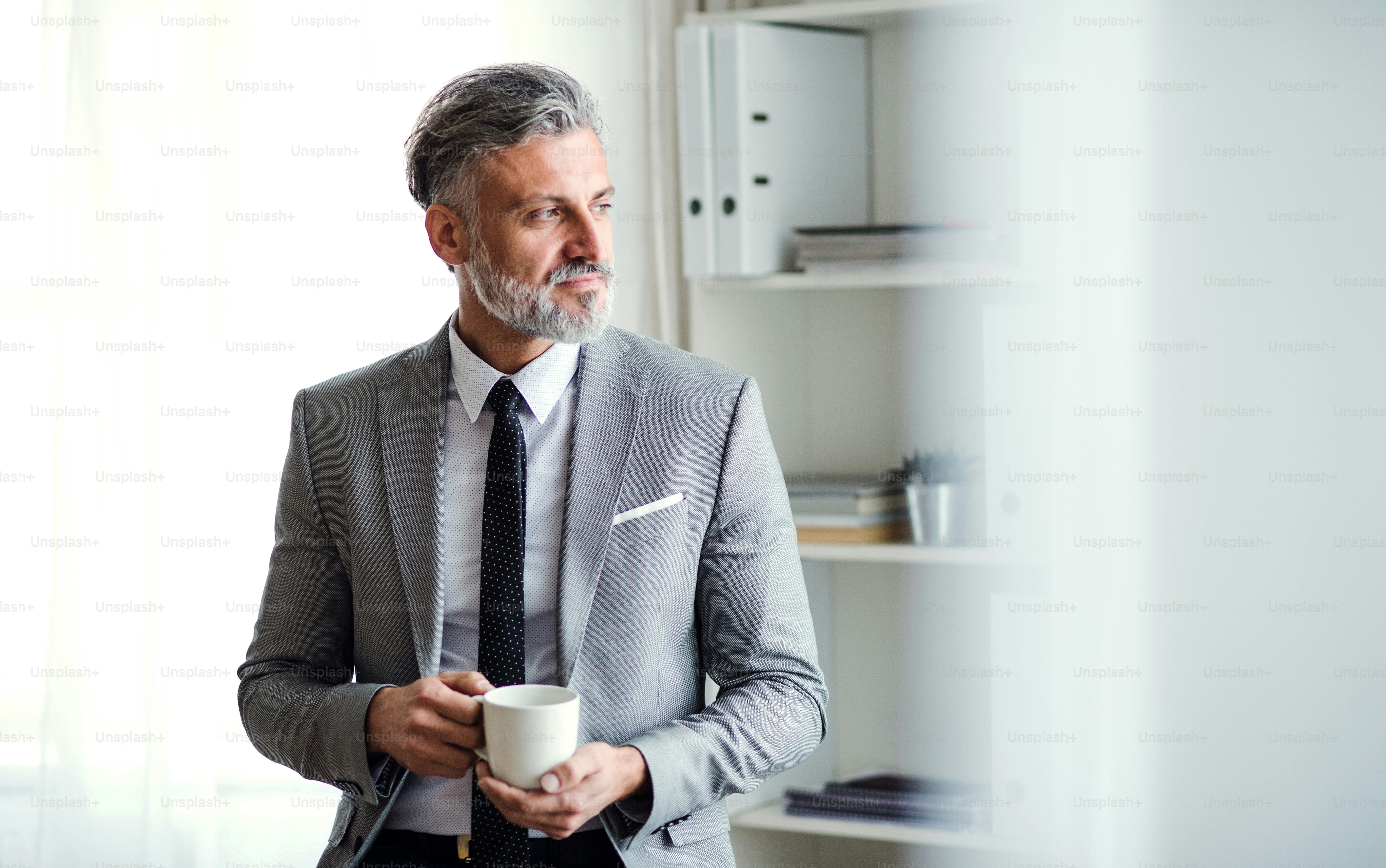 A serious mature businessman standing in an office, holding a cup of coffee. Copy space.