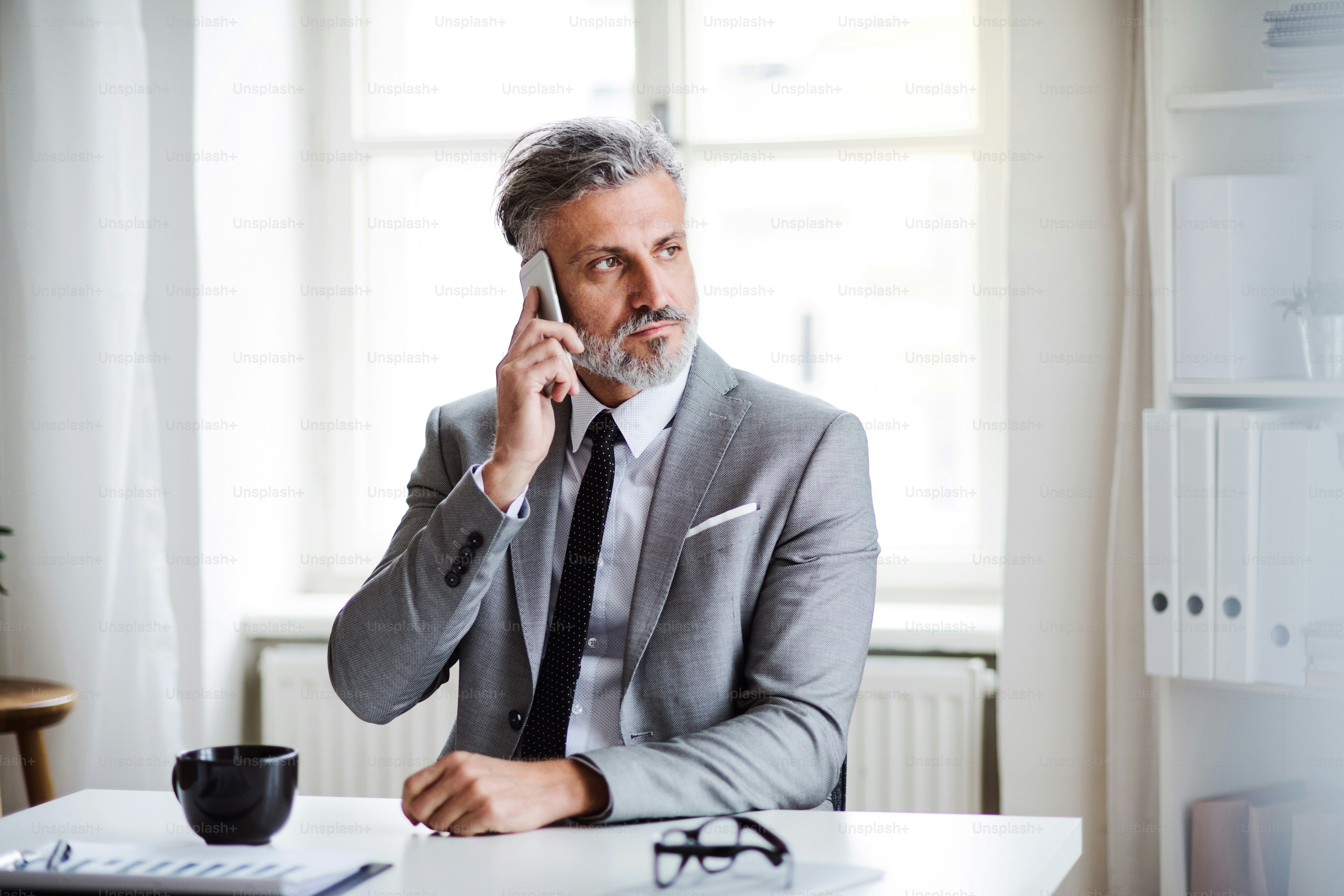 A serious mature businessman with smartphone sitting at the table in an office, making a phone call