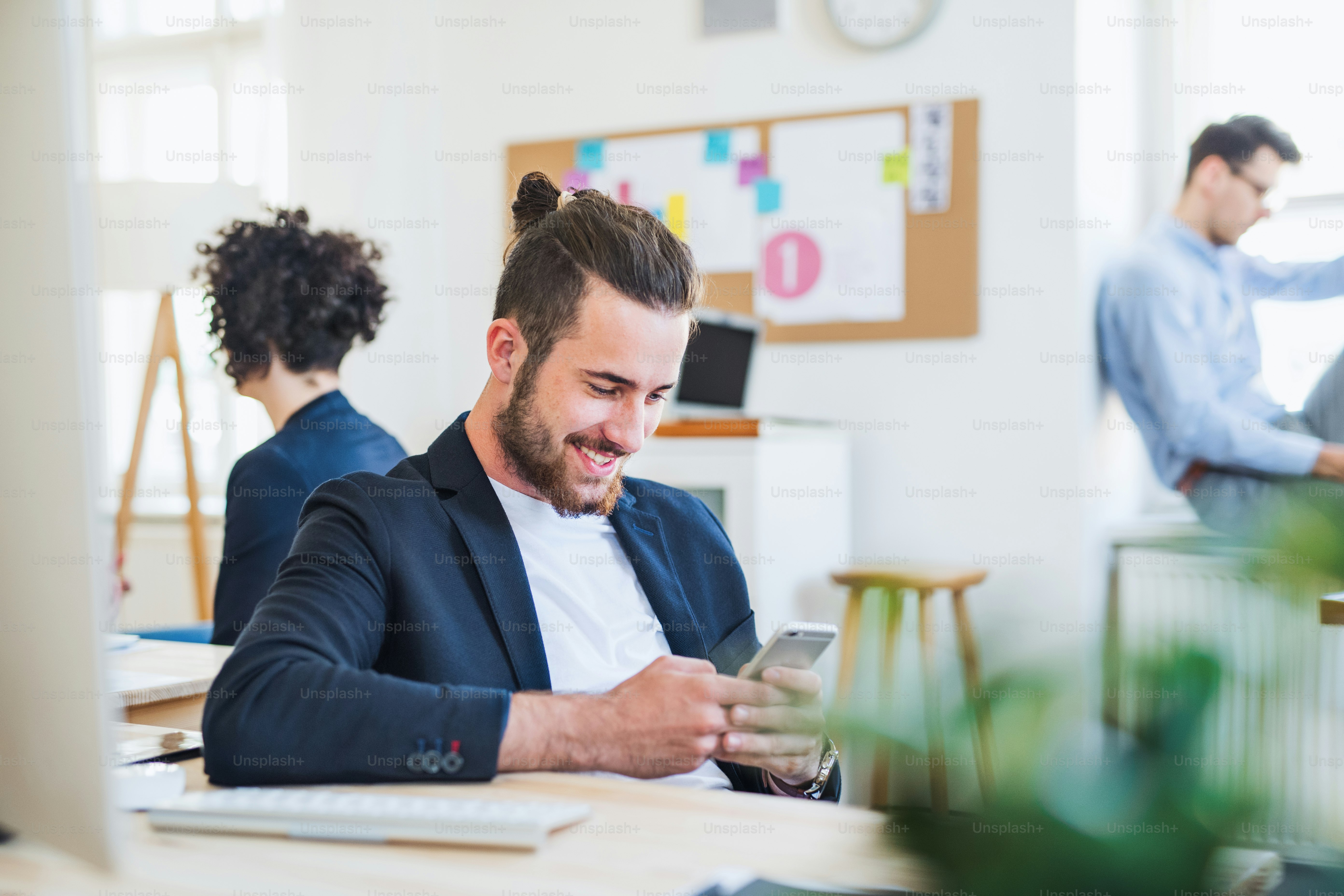 Potret pengusaha hipster muda dengan smartphone dan rekan-rekan di latar belakang di kantor modern.
