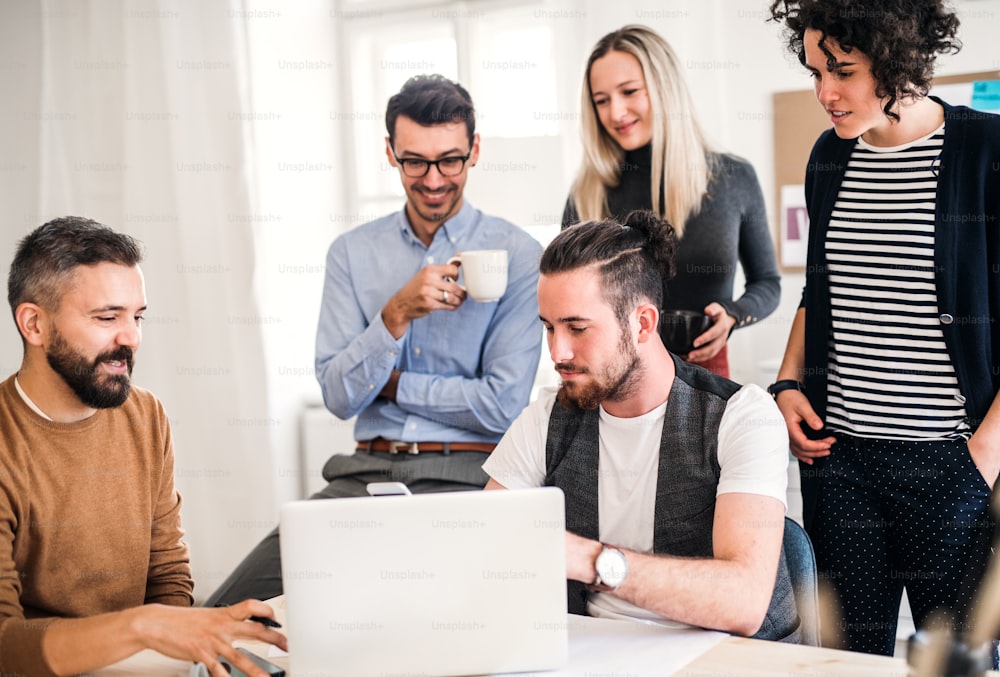 Grupo de empresários jovens e alegres com laptop sentado e em pé ao redor da mesa em um escritório moderno, tendo reunião.