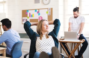 Portrait of young relaxed businesswoman with colleagues in a modern office, hands behind head and feet on desk.