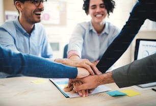 Grupo de jóvenes empresarios irreconocibles sentados alrededor de la mesa en una oficina moderna, apilando las manos.