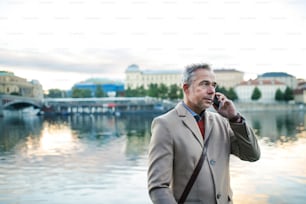 Mature handsome businessman with smartphone standing by river Vltava in Prague city, making a phone call. Copy space.