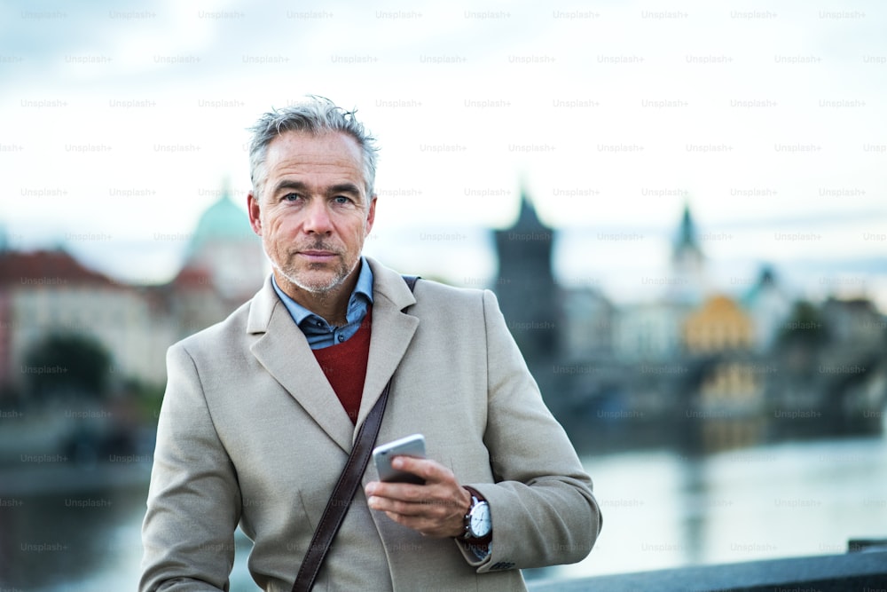 Mature handsome businessman with smartphone standing by river Vltava in Prague city. Copy space.