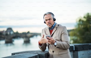 Mature handsome businessman with headphones and smartphone standing by river Vltava in Prague city, listening to music and singing. Copy space.