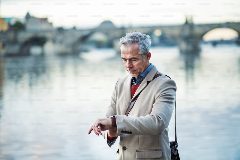 Mature handsome businessman standing by river Vltava in Prague city, checking the time.