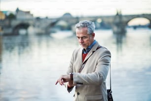 Mature handsome businessman standing by river Vltava in Prague city, checking the time.