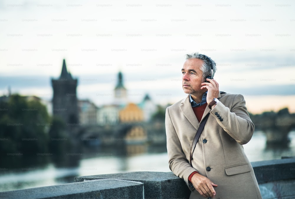 Hombre de negocios maduro y guapo con auriculares de pie junto al río Vltava en la ciudad de Praga, escuchando música. Espacio de copia.