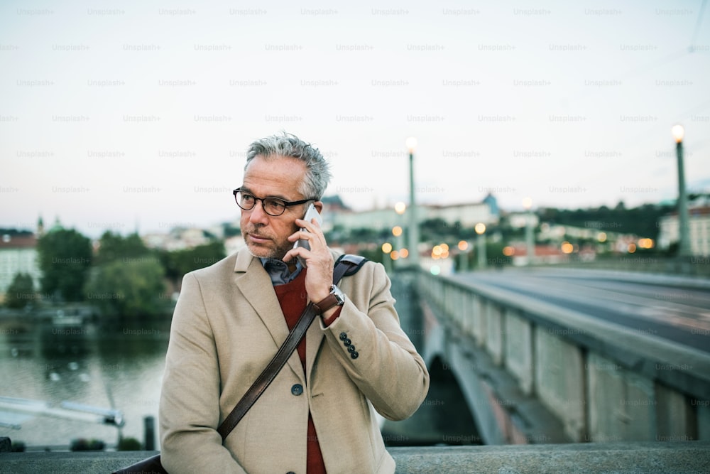 Mature handsome businessman with glasses and smartphone standing by river Vltava in Prague city, making a phone call. Copy space.