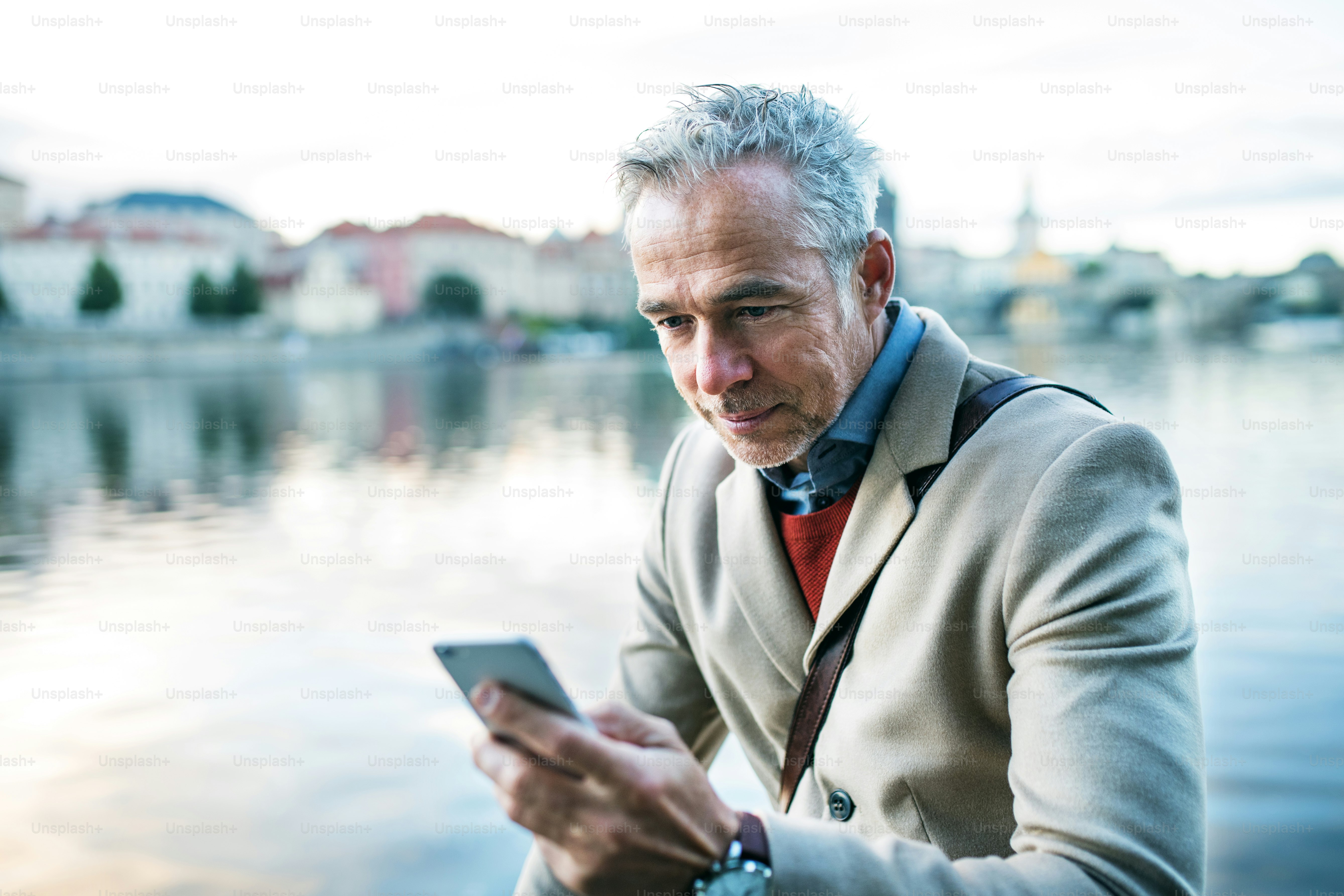 Mature handsome businessman with smartphone standing by river Vltava in Prague city. Copy space.