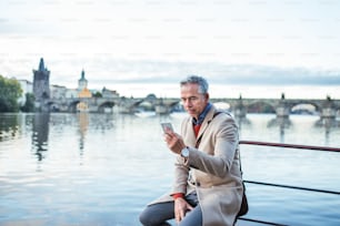 Mature handsome businessman with smartphone sitting by river Vltava in Prague city. Copy space.