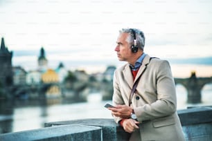 Mature handsome businessman with headphones and smartphone standing by river Vltava in Prague city, listening to music. Copy space.