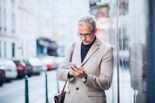 Mature businessman standing on a street in city, holding smartphone. Copy space.