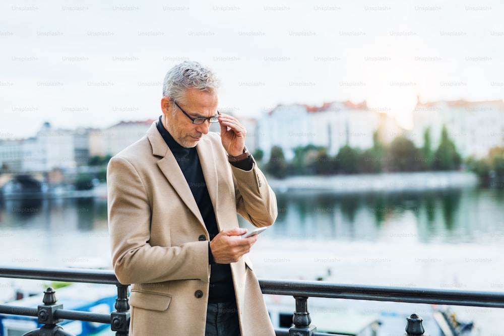 Hombre de negocios guapo maduro con teléfono inteligente de pie junto al río Vltava en la ciudad de Praga al atardecer, usando un teléfono inteligente.