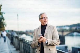 Mature handsome businessman with smartphone standing by river Vltava in Prague city at sunset, using smartphone.