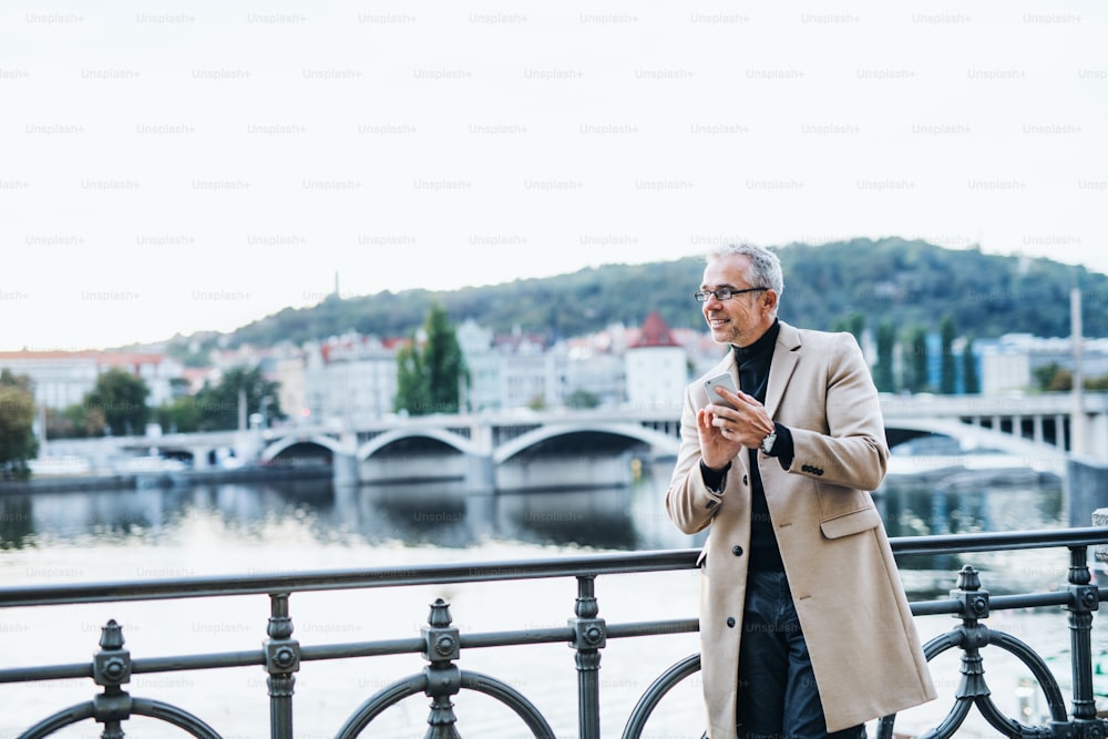 Mature handsome businessman with smartphone standing by river Vltava in Prague city at sunset, texting. Copy space.