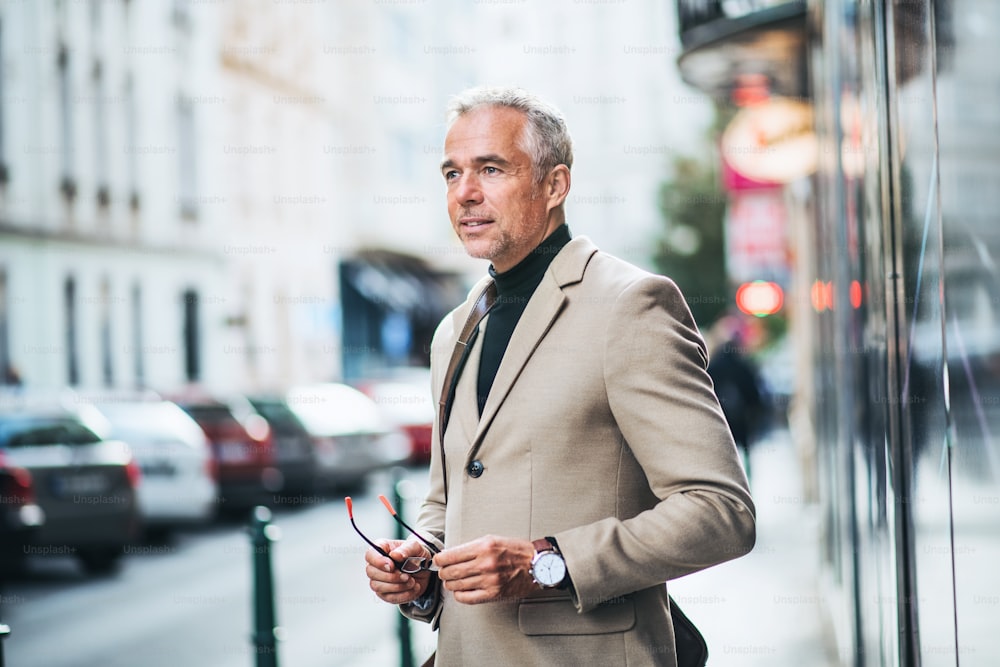 Mature businessman standing on a street in city, holding glasses. Copy space.