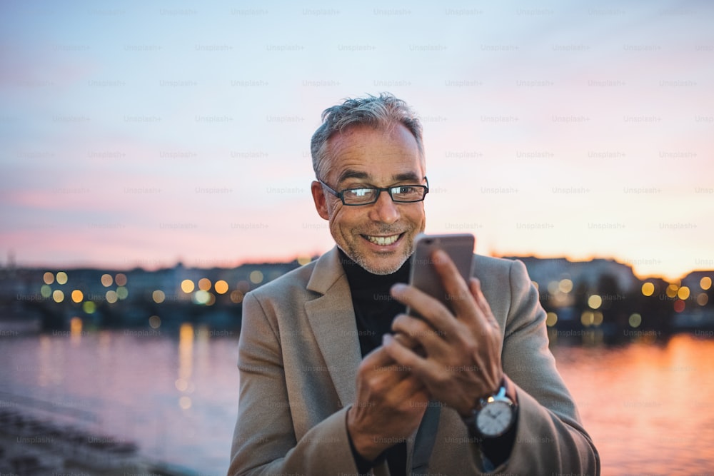 Mature handsome businessman with smartphone standing by river Vltava in Prague city at dusk time, taking selfie.