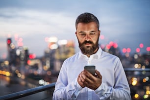 A portrait of businessman with smartphone standing against night London rooftop view panorama at sunset, text messaging. Copy space.