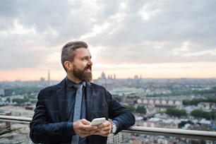 A portrait of businessman with smartphone standing against London rooftop view panorama, texting. Copy space.