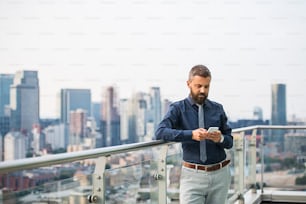 A portrait of businessman with smartphone standing against London rooftop view panorama, texting. Copy space.