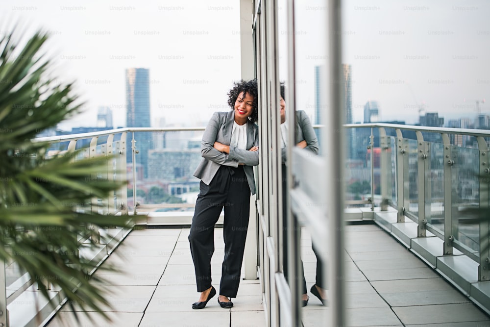 A portrait of black businesswoman standing on a terrace against London rooftop view panorama, arms crossed. Copy space.