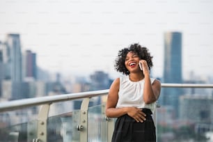 A portrait of happy black businesswoman standing against London rooftop view panorama, making a phone call. Copy space.