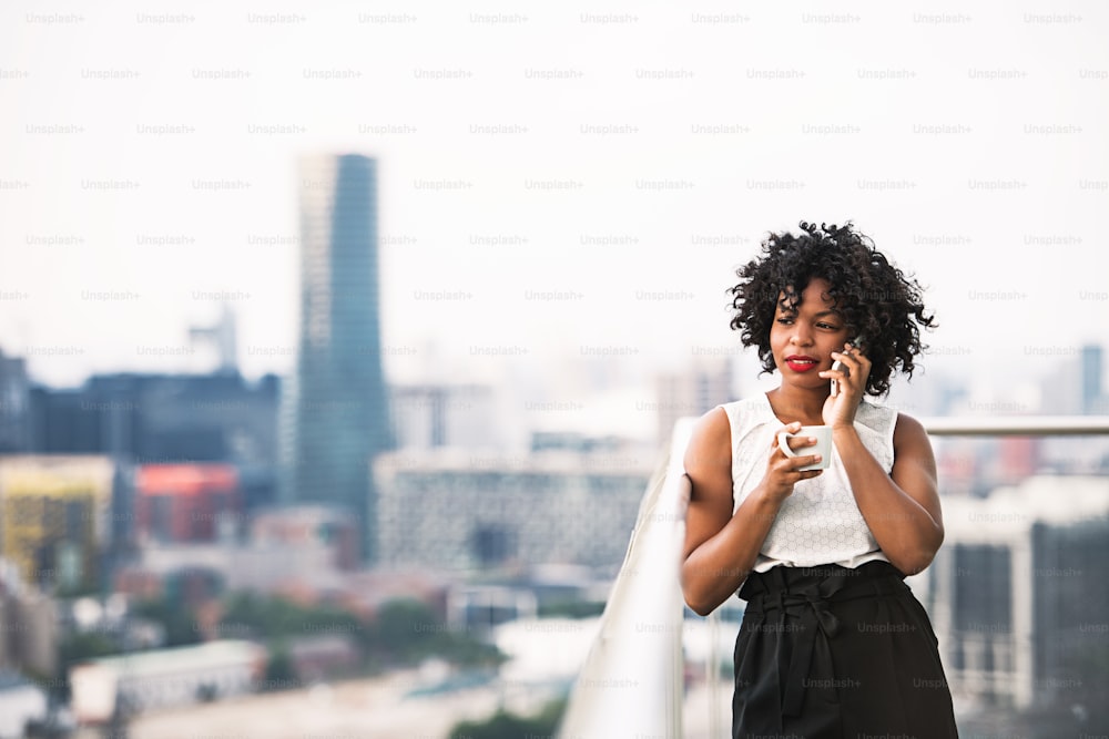 A portrait of black businesswoman with a cup of coffee standing against London rooftop view panorama, making a phone call. Copy space.