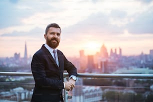 A portrait of businessman with jacket standing against London rooftop view panorama at sunset. Copy space.
