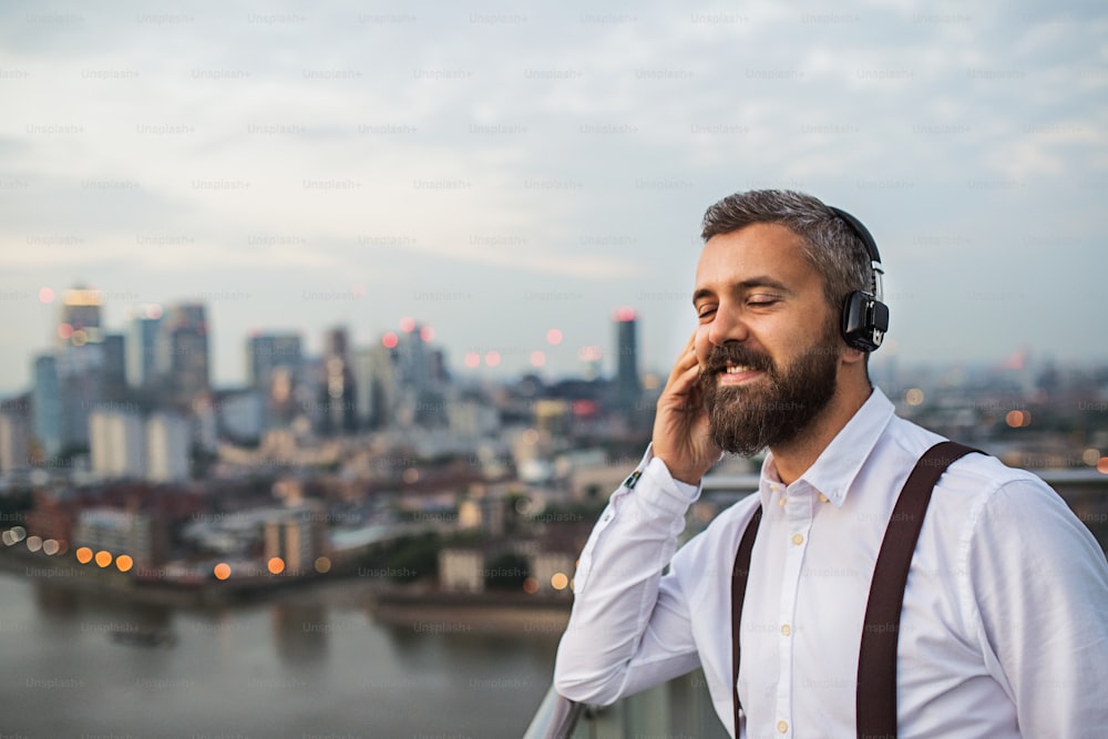 Un hombre de negocios con auriculares parado frente al panorama de Londres, escuchando música. Espacio de copia.
