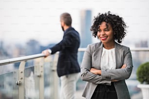 A portrait of black businesswoman standing with a colleague in the background on a terrace against London rooftop view panorama, arms corssed. Copy space.